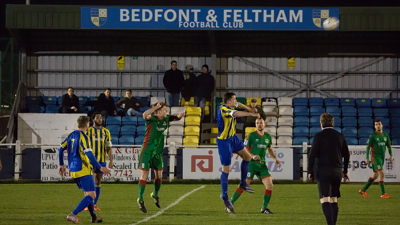 A floodlit evening non-league match, players contesting a header in front of a stand where fewer than a dozen spectators are sitting among rows of empty blue and yellow seats