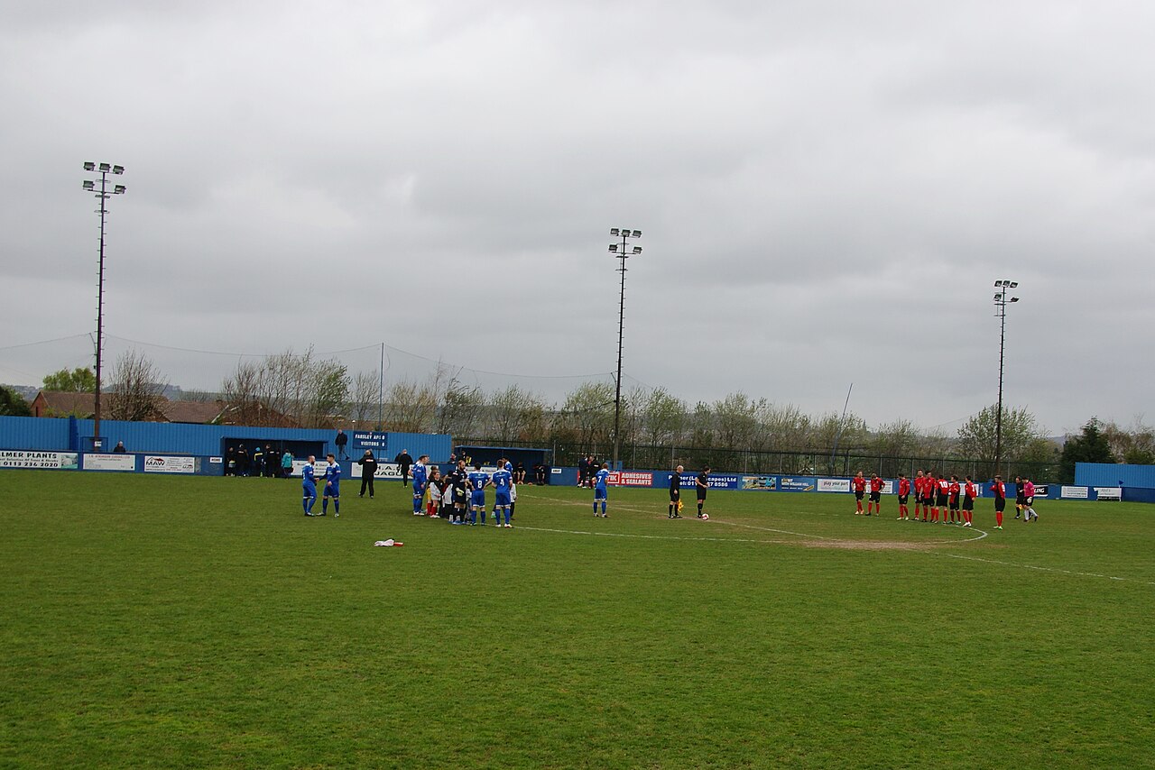 Two non-league teams lining up on a green pitch under a flat grey sky, with a low blue perimeter wall, three floodlight pylons and only a handful of people watching
